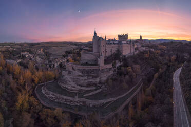 Aerial view of ancient castle Alcazar of Segovia Spain located on hill with trees next to highway with lamps against cloudy sky during twilight with bright moon - ADSF49185