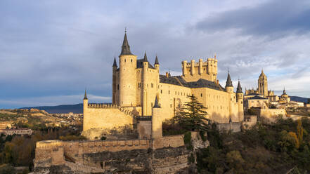 Amazing view of medieval castle Alcazar of Segovia Spain located on hill with green trees in sunny daylight against cloudy blue sky in evening time - ADSF49186