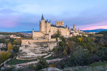 Top view of aerial view of medieval castle Alcazar of Segovia Spain located on hill with autumn trees next to meandering highway city buildings in sunny daylight - ADSF49187