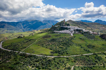 Blick auf ein grünes Tal mit einem kleinen Dorf und einem Bergrücken unter bewölktem Himmel bei Tag in Zahara de la Sierra in der Region Pueblos Blancos in Andalusien - ADSF49235