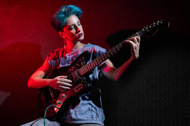 Thoughtful stylish young female musician in casual clothes with dyed hair and makeup passionately playing guitar while on dark red studio background - ADSF49245