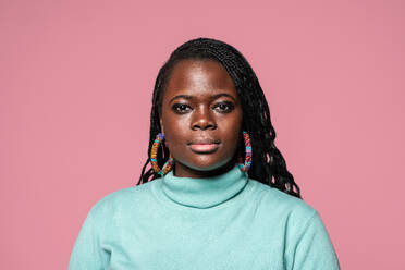 Portrait of an African woman with braided hair wearing colorful earrings and a turquoise turtleneck against a pink background - ADSF49257