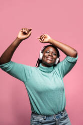 An African woman in a green top joyfully raising her arms while wearing white headphones against a pink background - ADSF49267