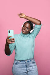A cheerful African woman with headphones dances while taking a selfie against a pink background - ADSF49278