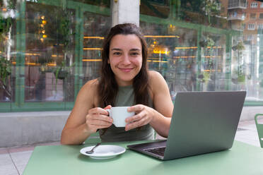 Young calm female freelancer sitting at table with laptop and drinking coffee while working on remote project in cafe terrace with blur background - ADSF49289