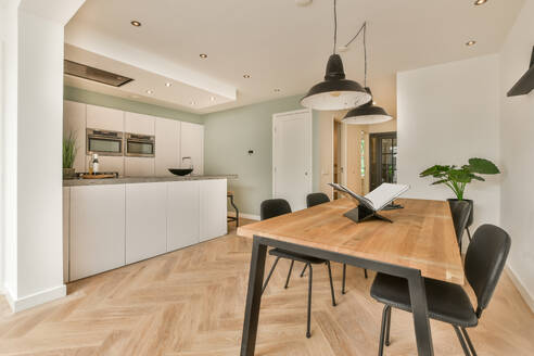 Book on wooden dining table with chairs and pendant lights in front of modern white kitchen at home - ADSF49352