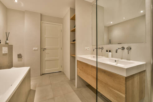 Ceramic bathtub with sink and mirror in illuminated bathroom at contemporary apartment with white walls - ADSF49354