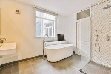 Ceramic bathtub with glass shower cabin and window in spacious luxurious bathroom at modern apartment - ADSF49360