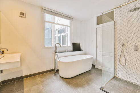 Ceramic bathtub with glass shower cabin and window in spacious luxurious bathroom at modern apartment - ADSF49360