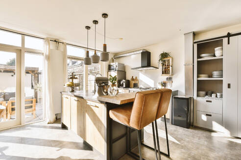 Brown empty chairs placed by island in open plan kitchen against bright window in modern apartment - ADSF49372