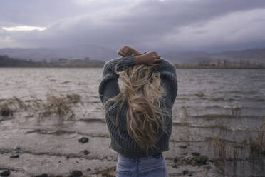 Blond woman with arms raised standing in front of lake at sunset - YBF00313