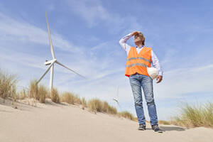 Engineer shielding eyes and standing near wind turbines on sunny day - PHDF00098