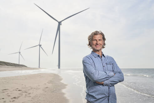 Lächelnder Geschäftsmann vor einer Windkraftanlage am Strand - PHDF00100