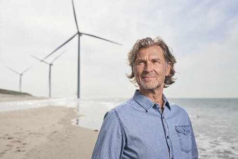 Lächelnder Geschäftsmann vor einer Windkraftanlage am Strand - PHDF00101