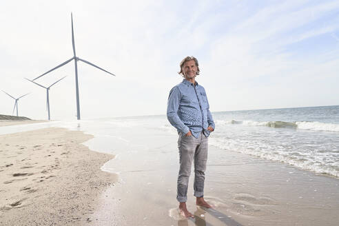 Ein lächelnder älterer Geschäftsmann steht vor Windkraftanlagen am Strand - PHDF00103