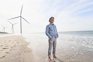 Älterer Geschäftsmann steht vor Windrädern am Strand - PHDF00104