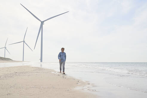 Geschäftsmann geht vor Windkraftanlagen am Strand spazieren - PHDF00107