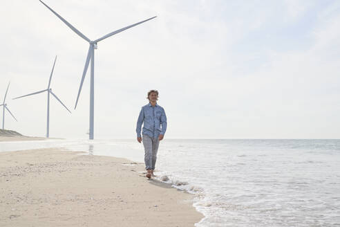 Älterer Geschäftsmann geht vor Windkraftanlagen am Strand spazieren - PHDF00108