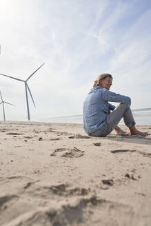 Mann sitzt in der Nähe von Windrädern am Strand unter Himmel - PHDF00110