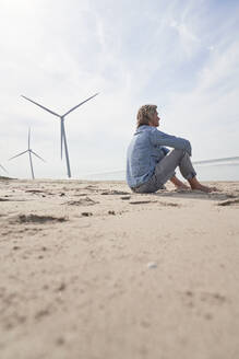 Älterer Mann sitzt in der Nähe von Windrädern am Strand unter dem Himmel - PHDF00111