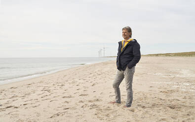 Älterer Mann steht mit Händen in den Taschen am Strand - PHDF00115