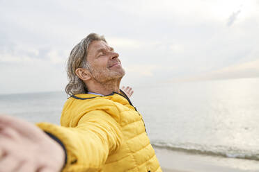 Smiling senior man with arms outstretched standing at beach - PHDF00130