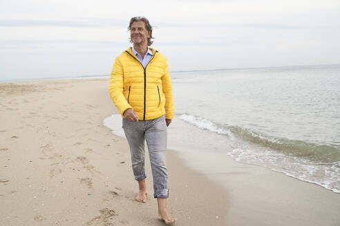 Lächelnder Mann, der an der Küste in der Nähe des Meeres am Strand spazieren geht - PHDF00139