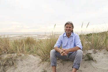 Lächelnder älterer Mann sitzt auf Sand am Strand - PHDF00142