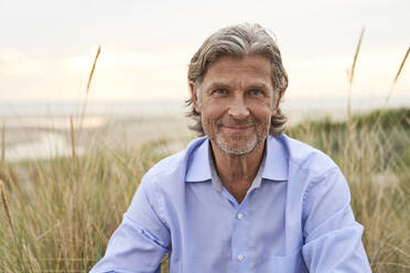 Lächelnder Mann sitzt in der Nähe von Gras am Strand - PHDF00143