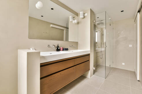 Mirror over ceramic sink and wooden cabinets with glass shower cabin in modern bathroom at home - ADSF49403