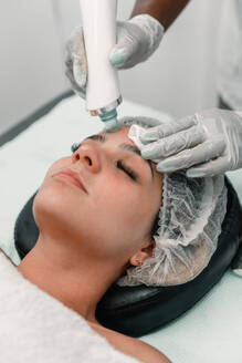 High angle of crop ethnic beautician in gloves applying toning treatment with cream and cotton pad on face of young woman resting on back in spa - ADSF49406