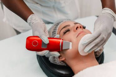 High angle of crop beautician in gloves using skin scrubber and cotton on face of young female client lying on back and resting head on pillow in spa - ADSF49407