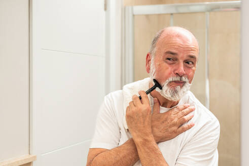 Content elderly gray haired male shaving beard with razor in light bathroom during morning hygienic procedure - ADSF49410