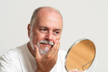 Elderly bearded man holding round mirror and touching face on white backdrop during daily beauty treatment - ADSF49412