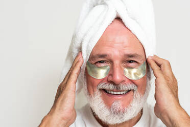 Glad senior man with towel on head looking at camera and applying eye patches during skincare routine against white background - ADSF49413