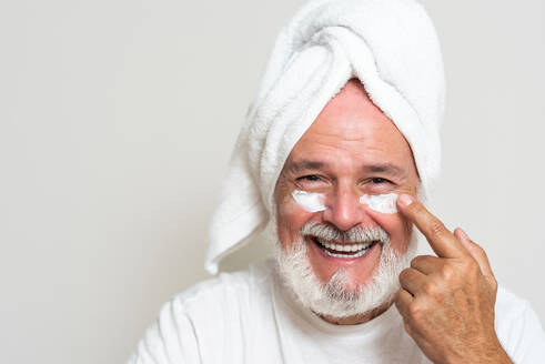 Happy senior bearded man with towel on head applying moisturizing cream on face while looking at camera - ADSF49414