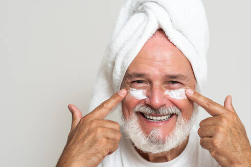 Happy senior bearded man with towel on head applying moisturizing cream on face while looking at camera - ADSF49415