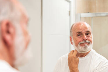 Positive elderly man shaving gray beard with sharp blade while standing in bathroom looking at himself in mirror during morning routine - ADSF49417