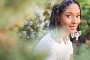 Side view of young African American female with braids and sitting in park against blooming flowers on background of nature and enjoying time outside - ADSF49418