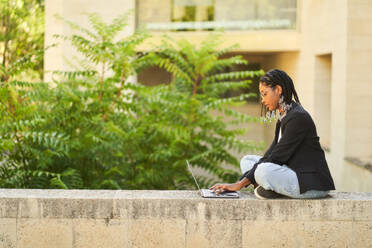 Side view of African American businesswoman sitting on stone border and working remotely in front of building with laptop while browsing internet - ADSF49421