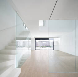 Bright and minimalist interior of a modern home featuring a white staircase with clear glass railings, leading to a spacious room with large sliding doors opening to a terrace. - ADSF49449