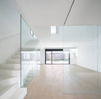 Bright and minimalist interior of a modern home featuring a white staircase with clear glass railings, leading to a spacious room with large sliding doors opening to a terrace. - ADSF49449