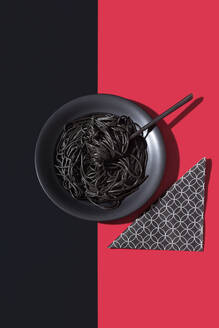 Top view of black spaghetti in a bowl with fork placed on black and red background near napkin - ADSF49458