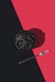 Top view of black spaghetti in a bowl placed on black and red background near fork - ADSF49460