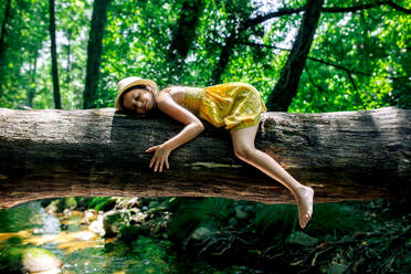 Side view of preteen girl in hat and dress with closed eyes lying on tree trunk in summer day against green forest in Gorge Africa - ADSF49461