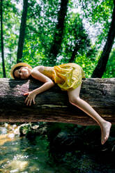Side view of smiling preteen girl in hat and dress with closed eyes lying on tree trunk in summer day against green forest in Gorge Africa - ADSF49464
