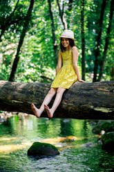 Smiling preteen girl wearing hat and dress looking at barefoot while sitting on tree trunk in summer day against green forest in Gorge Africa - ADSF49470