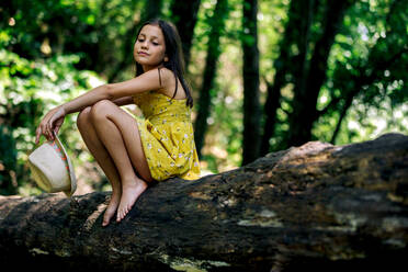 Side view of pensive preteen girl in dress holding hat while sitting on tree trunk in summer day against green forest in Gorge Africa - ADSF49471