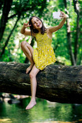 Smiling preteen girl wearing dress sitting on tree trunk while taking selfie with smartphone in summer day against green forest in Gorge Africa - ADSF49473