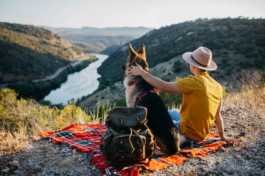 Back view of anonymous male hiker in yellowt shirt sitting on green hill with adorable dog in red collar and admiring picturesque scenery - ADSF49478